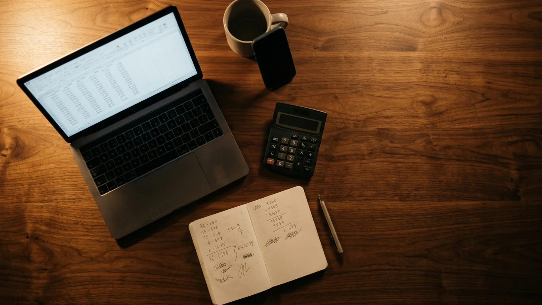 A desk at night with a laptop showing a spreadsheet of revenue rows, a calculator, a notebook of hand-written figures and a phone propped beside it, lit by a single warm lamp.